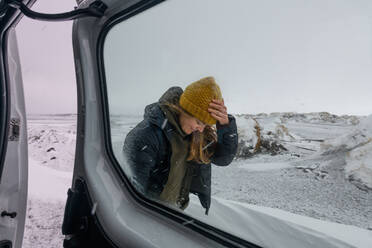 Eine Frau mit Wollmütze und Jacke steht durch das Fenster eines Wohnmobils in einer verschneiten Landschaft: Wintersurfen. - ISF24071