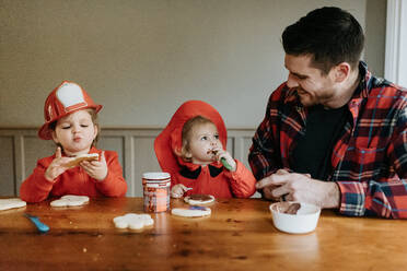 Zwei als Feuerwehrmänner verkleidete Kinder und ihr Vater sitzen an einem Tisch und verzieren Kekse mit Schokolade und Streuseln. - ISF24082