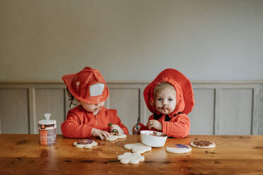 Zwei als Feuerwehrmänner verkleidete Kinder sitzen an einem Tisch und verzieren Kekse mit Schokolade und Streuseln. - ISF24083