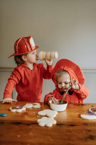 Zwei als Feuerwehrmänner verkleidete Kinder sitzen an einem Tisch und verzieren Kekse mit Schokolade und Streuseln., lizenzfreies Stockfoto