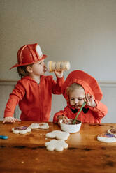 Zwei als Feuerwehrmänner verkleidete Kinder sitzen an einem Tisch und verzieren Kekse mit Schokolade und Streuseln. - ISF24084