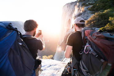 Rückansicht von zwei Männern, die mit einem Mobiltelefon Fotos von Felsen und einem Sonnenuntergang machen. - ISF24099