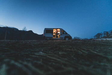 Campervan silhouetted on a hill in the evening with the lights on inside and a man standing by the open side door. - ISF24103
