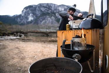 Ein Mann kocht auf einem Grill, der auf der Rückseite des Wohnmobils in einer winterlichen Landschaft aufgebaut ist. - ISF24118