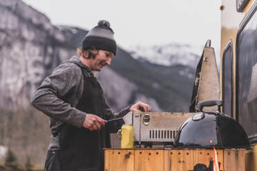 Ein Mann kocht auf einem Grill, der auf der Rückseite des Wohnmobils in einer winterlichen Landschaft aufgebaut ist. - ISF24119