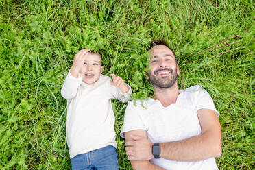 Blick von oben auf einen lächelnden, bärtigen Mann und einen kleinen Jungen, die im Gras liegen. - ISF24161