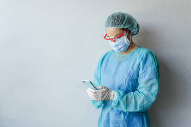 Mature female doctor in medical scrubs using smart phone while standing against white wall at clinic - JCMF00732