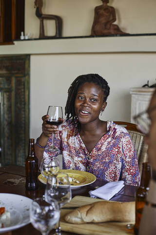 Lächelnde junge Frau mit Weinglas in der Hand, die während des Mittagessens am Esstisch sitzt, lizenzfreies Stockfoto