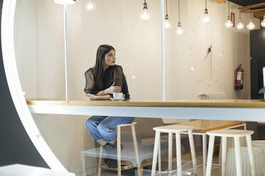 Schöne Frau in voller Länge, die wegschaut, während sie in einem beleuchteten Cafe sitzt und wartet - OCAF00462