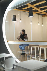 Lächelnde junge Frau, die in einem beleuchteten Café sitzt und mit ihrem Handy telefoniert - OCAF00463
