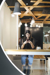 Full length of confident young woman using mobile phone while sitting at illuminated cafeteria - OCAF00465