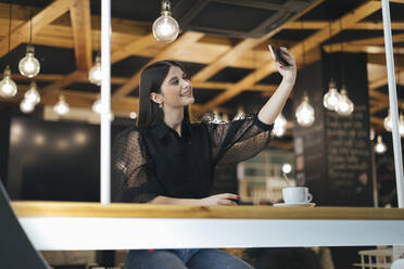 Glückliche junge Frau, die ein Selfie mit ihrem Smartphone macht, während sie in einem Café sitzt - OCAF00468
