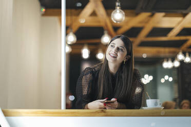 Happy young woman looking away while sitting at illuminated cafe - OCAF00471