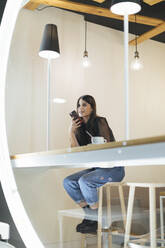 Full length of beautiful woman sitting with smart phone and coffee at cafe - OCAF00476