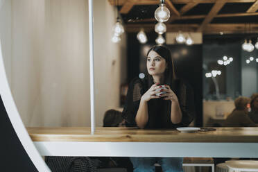 Beautiful young woman looking away while sitting with coffee cup thinking at cafe - OCAF00479