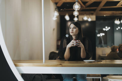 Schöne junge Frau schaut weg, während sie mit einer Kaffeetasse im Café sitzt und nachdenkt - OCAF00479
