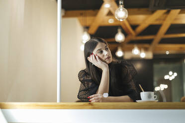 Thoughtful young woman sitting at table in illuminated coffee shop - OCAF00480