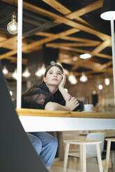 Thoughtful young woman leaning on table while looking away at illuminated cafeteria - OCAF00481