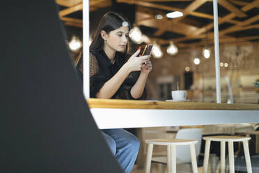 Schöne junge Frau, die ein Mobiltelefon benutzt, während sie in einem beleuchteten Café sitzt - OCAF00482