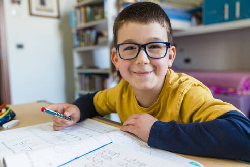 Smiling cute boy sitting with book at desk during COVID-19 homeschooling - MGIF00940
