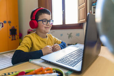 Lächelnder männlicher Schüler, der während des Hausunterrichts über Kopfhörer zuhört und auf seinen Laptop schaut - MGIF00951