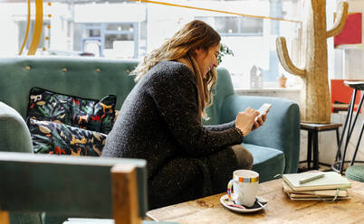 Lachende Frau mit Smartphone auf der Couch in einem Café sitzend - MGOF04255