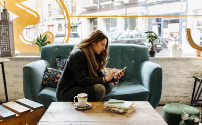 Woman using smartphone sitting on couch in coffee shop - MGOF04256