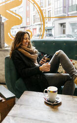 Lachende Frau mit Smartphone auf der Couch in einem Café sitzend - MGOF04257