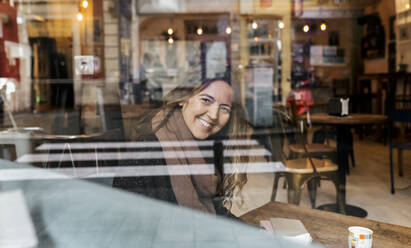 Lächelnde Frau, die durch die Fensterscheibe eines Cafés schaut - MGOF04260
