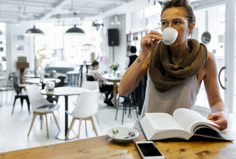 Frau liest in einem Café ein Buch und trinkt Kaffee - MGOF04280