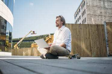 Businessman with laptop sitting on skateboard at construction site in the city - JOSEF00556