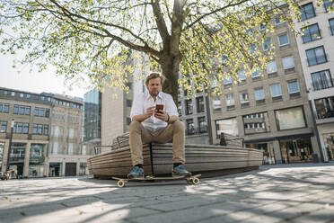 Geschäftsmann mit Skateboard sitzt auf einer Bank in der Stadt und prüft sein Smartphone - JOSEF00561