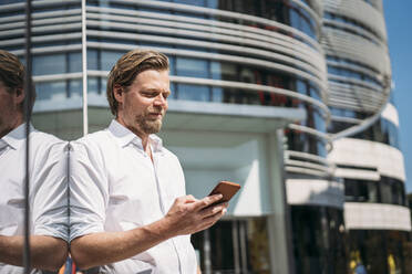 Businessman in the city leaning against glass front checking smartphone - JOSEF00572