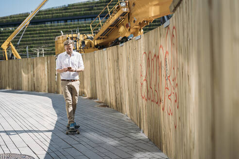 Geschäftsmann fährt Skateboard und benutzt sein Smartphone auf einer Baustelle in der Stadt - JOSEF00581
