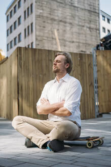 Geschäftsmann sitzt auf Skateboard auf einer Baustelle in der Stadt mit geschlossenen Augen - JOSEF00582