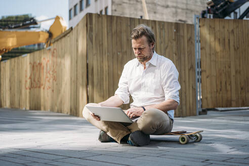 Geschäftsmann mit Laptop auf dem Skateboard auf einer Baustelle in der Stadt - JOSEF00583