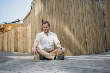 Businessman sitting on the ground at construction site with wooden house model - JOSEF00586