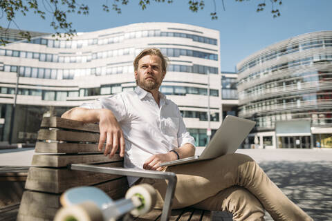 Geschäftsmann sitzt auf einer Bank in der Stadt und benutzt einen Laptop, lizenzfreies Stockfoto