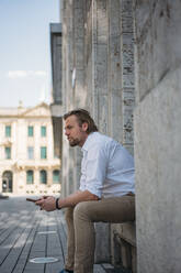Businessman sitting in niche of a building holding smartphone - JOSEF00630
