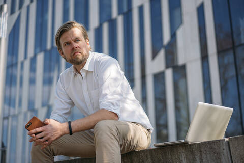 Businessman with laptop and smartphone having a break in the city sitting on a wall stock photo