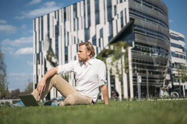 Businessman with laptop sitting in grass in the city - JOSEF00652