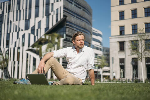 Geschäftsmann mit Laptop im Gras sitzend in der Stadt, lizenzfreies Stockfoto