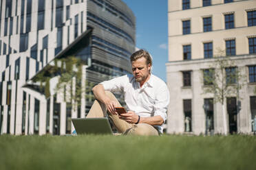 Businessman with laptop sitting in grass in the city using smartphone - JOSEF00654