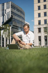 Businessman with laptop sitting in grass in the city using smartphone - JOSEF00655