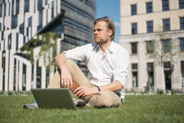 Businessman with laptop sitting in grass in the city - JOSEF00656