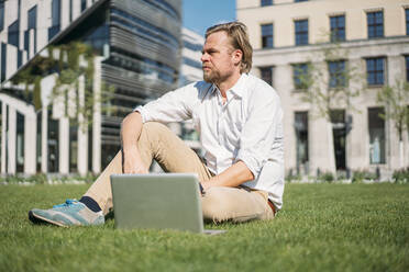 Businessman with laptop sitting in grass in the city - JOSEF00658