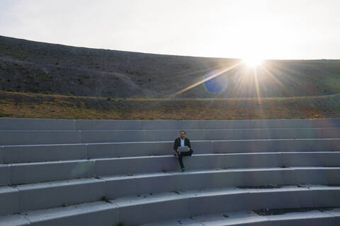 Geschäftsmann sitzt mit Laptop bei BergArena Amphitheater Schritte gegen den Himmel bei Sonnenuntergang, lizenzfreies Stockfoto