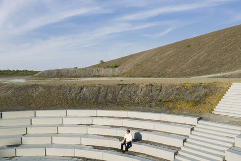 Männlicher Berufstätiger auf den Stufen eines Amphitheaters gegen den Himmel an einem sonnigen Tag - MOEF02874