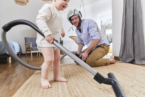 Mann mit Sturzhelm schaut auf seinen kleinen Sohn, der den Teppich im Wohnzimmer mit dem Staubsauger reinigt - FSF01074