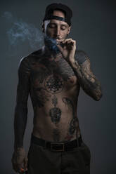 Porträt eines tätowierten Mannes vor grauem Hintergrund, der einen Joint raucht - AMUF00108